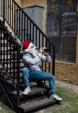 Evsiz Noel Baba şarap içiyor ve çöp kutularını karıştırıyor.