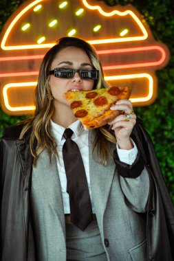 Genç iş kadını sokakta neon ışıklarının önünde pizza yiyor.