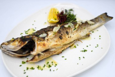Limonlu ızgara balık ve bir restoranda beyaz bir tabakta otlar.