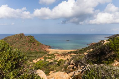 Menorca 'daki boş sahil Cala del Pilar, kristal berrak denizli kırmızı dağ ve turuncu kum.