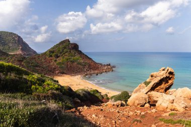 Menorca 'daki boş sahil Cala del Pilar, kristal berrak denizli kırmızı dağ ve turuncu kum.