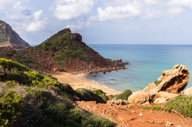 Menorca 'daki boş sahil Cala del Pilar, kristal berrak denizli kırmızı dağ ve turuncu kum.