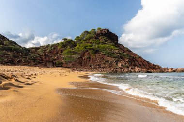 Menorca 'daki boş sahil Cala del Pilar, kristal berrak denizli kırmızı dağ ve turuncu kum.