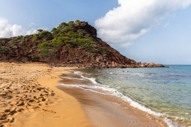 Menorca 'daki boş sahil Cala del Pilar, kristal berrak denizli kırmızı dağ ve turuncu kum.