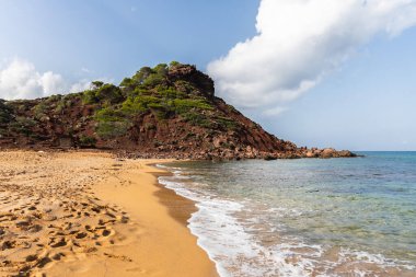 Menorca 'daki boş sahil Cala del Pilar, kristal berrak denizli kırmızı dağ ve turuncu kum.