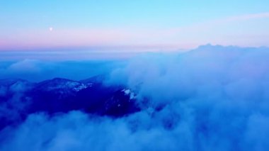 Güneş doğmadan önceki mavi saatte bulutların ve dağların üzerinden sabah uçuşunun alçak hava görüntüsü. Ufuktaki ay. Geriye doğru hareket. Yüksek kalite 4k görüntü