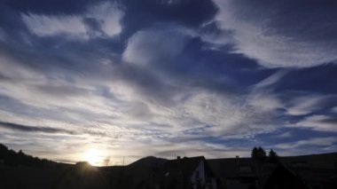 Köydeki evlerin çatıları üzerindeki zaman atlamalı bulutlar. Günbatımı, pastel renkli bulutlar, sabahtan akşama kadar. Yüksek kalite 4k görüntü