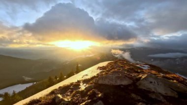 Kışın rüzgarlı dağ manzarası, kar ve çimlerle kaplı tepe sırtı, kalın bulutlar, uzaktaki kayaların üzerinde güneş ışığı parlıyor. Yüksek kalite 4k görüntü