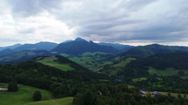 Bir yaz günü, hava hipersüratli drone videosu geniş yeşil bir arazide yavaşça yana kayar. Sahnede kayan tepeler, sık ormanlar, bir muhafazadaki koyunlar, küçük bir köy ve