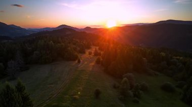 Dinamik bir hiperhız videosu canlı bir yaz günbatımında bir dağ silsilesinin havadan görüntüsünü yakalar. Güneş tepelerin ardında batıyor, yeşil manzaranın üzerine sıcak bir altın parıltısı saçıyor ve