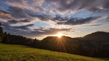 Bir zaman dilimi videosu, tepelik bir arazide güzel bir gün batımı yakalar. Güneş ufuktan iniyor, dağların kenarındaki yeşil çayırlar ve ormanı aydınlatıyor, gökyüzü ise...