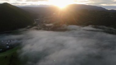 Bir hava görüntüsü, bir dağ vadisini dolduran alçak bir sis bulutunu ve sisin altında kısmen görünen küçük bir köyü yakalıyor. Karanlık dağlar sahneyi oluşturur ve yükselen güneş güçlü bir güneş yaratır.
