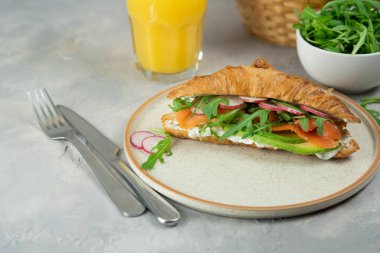Croissant sandwich with cream cheese, salmon and avocado on a plate, gray background, close-up. Healthy breakfast concept.