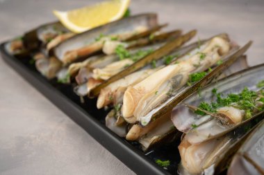 Dish of razor clam grilled with garlic, parsley, olive oil and lemon slice on gray stone table.