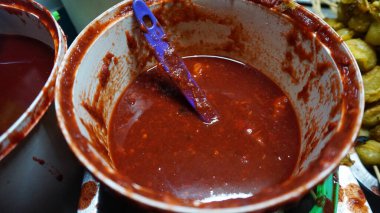 Kırmızı biber sosuyla dolu, içinde mor bir kaşık olan bir konteynırın yakın çekimi. Sos yoğun ve baharatlı görünüyor, görünen acı biber tohumlarıyla. Kapta kısmen sos lekesi var..