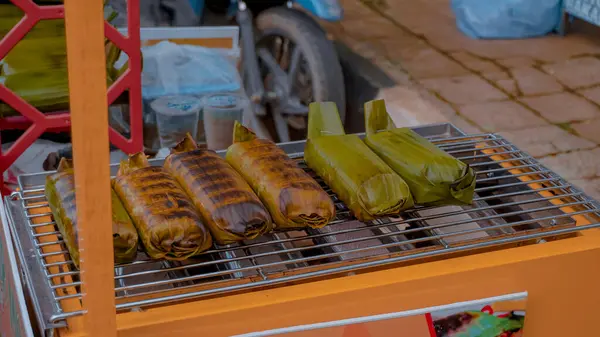 Muz yapraklarına sarılı çeşitli ızgara aperatifler, sokak yemekçisinin standında sergileniyor, geleneksel mutfak ve açık hava yemeklerinin özünü yakalıyor.