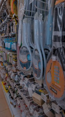 Bir mağaza koridorunda, raflarda düzgünce düzenlenmiş elektrikli sinek sinekleri, temizlik malzemeleri ve diğer aletler de dahil olmak üzere çeşitli ev eşyaları sergilenmektedir..