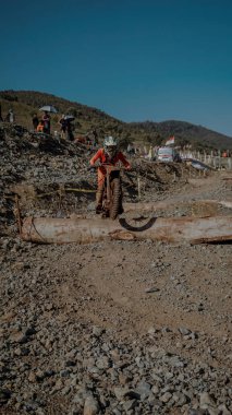 Bir motokros sürücüsü, açık mavi gökyüzü altında kayalık bir arazide engel parkurunda yol alıyor..