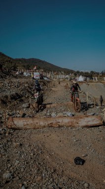 İki motosikletçi büyük bir kütük de dahil olmak üzere kayalık bir off-road yolunda engellerle geziniyor. Arka planda bir tepe manzarası ve olayı izleyen izleyiciler yer alıyor..