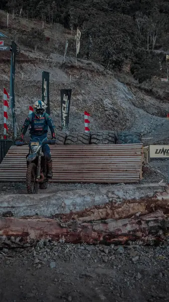 Bir motokros sürücüsü zorlu bir off-road pistinde kütükler ve lastikler de dahil olmak üzere engebeli bir çevrede geziniyor. Sürücü koruyucu giysi ve kask takıyor..