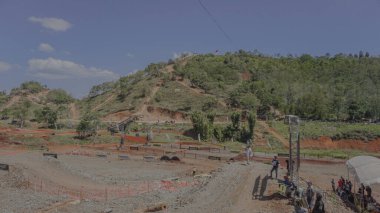 Tepe manzaralı bir motokros pisti. İzleyiciler olayı izliyor. Pist, açık mavi gökyüzünün altında yeşil ve tepelerle çevrili..