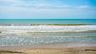 Açık mavi gökyüzünün altında kıyıya vuran yumuşak dalgaların olduğu sakin bir sahil sahnesi. Ufuk uzak teknelerle dolu, sakin bir atmosfer yaratıyor..