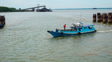 A vibrant blue fishing boat navigates a calm river with docks and industrial structures framing the scene, showcasing maritime livelihood and the coexistence of nature with human activity clipart