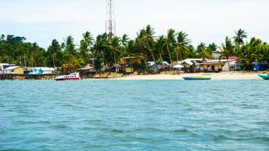 Tropikal bir sahil manzarası, palmiye ağaçları, sazlıklar üzerindeki renkli evler ve suya demirlenmiş tekneler. Kıyı şeridi kumlu ve arka planda bir telekomünikasyon kulesi görünüyor..