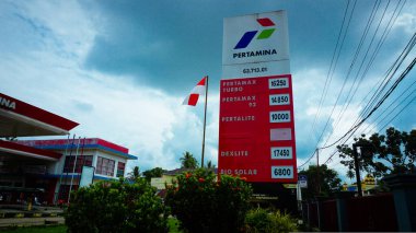 Bulutlu bir gökyüzünün altında yakıt fiyatlarını gösteren bir Pertamina benzin istasyonu tabelası..