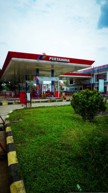 Yeşil ve bulutlu gökyüzü ile çevrili kırmızı ve beyaz bir gölgeliği olan bir Pertamina benzin istasyonu..