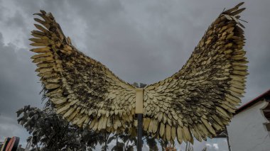 Ağaçlarla çevrili bulutlu bir gökyüzüne karşı büyük altın kanatlı heykel..