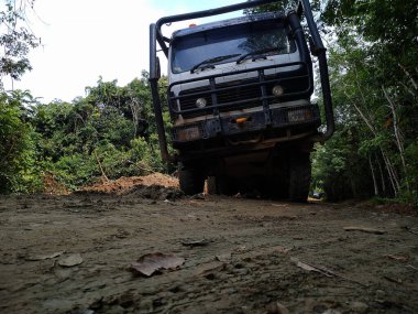 Çamurlu bir yolda, yemyeşil bir yolda büyük siyah bir kamyon. Kamyon yere yakın konumlanmış, dayanıklı lastiklerini ve dayanıklı yapısını gösteriyor. Arka planda, uzak bir bölgeyi gösteren sık ağaçlar ve yapraklar yer almaktadır..
