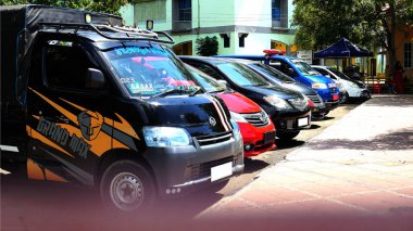 Sokakta park etmiş bir sürü araç var. Logolu siyah bir minibüs, kırmızı bir araba ve mavi bir polis aracı da dahil. Arka planda ağaçlar ve binalar olan şehir merkezi..