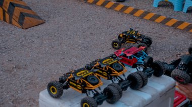 Toprak yolda uzaktan kumandalı arabaların olduğu bir sahne. Çeşitli modeller, renkli tasarımları ve sert lastikleri ile sergileniyor. Çevrede rampa ve bariyer gibi engeller var.