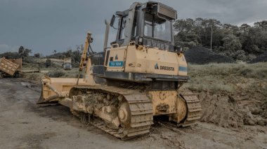 İnşaat alanında büyük sarı bir buldozer. Etrafı toprak ve bitki örtüsüyle çevrili. Buldozerde izler ve ön bıçak var. Çamurlu bir arazide konumlanmış..