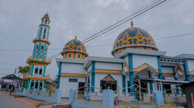 Uzun minaresi ve iki kubbesi olan renkli bir cami. Karmaşık geometrik desenleri olan, bulutlu bir gökyüzüne karşı kurulmuş.
