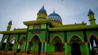 Mavi kubbeli, karmaşık desenli, birden fazla kemer ve açık gökyüzü arkaplanlı canlı yeşil bir cami..