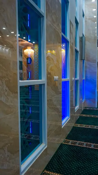 Zarif mermer duvarlar, mavi ışıklandırma ve dekoratif pencerelere sahip modern bir cami manzarası. Halı, karmaşık desenlerle yeşildir. Huzurlu bir atmosfer yaratır..