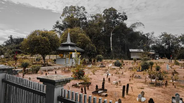 Çok sayıda mezar taşı ve yemyeşil yeşillik ile çevrili merkezi pagoda benzeri bir yapıya sahip huzurlu bir mezarlık sahnesi. Atmosfer sakin ve yansıtıcı, bulutlu bir gökyüzü var..