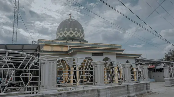 Kendine özgü kubbesi ve karmaşık mimari detayları olan bir cami. Etrafı çitlerle çevrili. Gökyüzü bulutlu, sahneye dramatik bir atmosfer ekliyor..