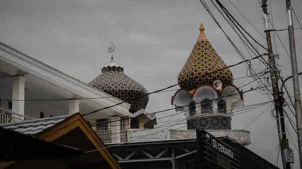 Bulutlu bir gökyüzünün altında, elektrik hatlarıyla çevrili eşsiz desenli kubbeleri olan bir cami.