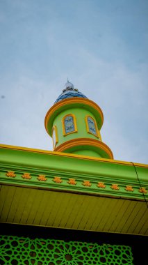 Mavi kubbeli yeşil ve sarı bir kule ve bulutlu gökyüzüne karşı dekoratif elementler içeren renkli bir mimari detay..