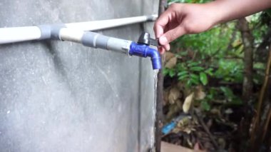 Gri bir PVC borusuna bağlı mavi plastik bir musluk ve içinden su akıyor. Arka planda yeşil yapraklar var. Açık hava ortamını gösteriyor..