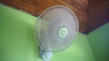 Daire şeklinde metal ızgaralı, yeşil duvarlı ve ahşap tavanlı, elektrikli bir vantilatör..