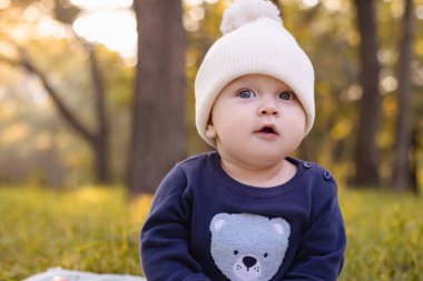 Örgü şapkalı bebek sonbahar ağaçlarının arka planında çimlerin üzerinde oturuyor. Çocuk kıyafetleri. Dışarı yürüyün. Yüksek kalite fotoğraf