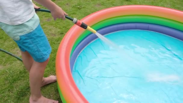少年は膨脹可能な多色のプールで水を集める 少年が持っていたホースから水が流れてくる 子供のための夏のレクリエーションとエンターテイメントのアイデア 高品質4K映像 — ストック動画