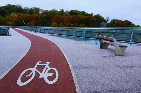 stock image Scenic autumn landscape view of winding red color bike lane with white bicycle sign on the New Pedestrian Bridge in Kyiv, Ukraine. Beautiful autumn colored trees in the background.