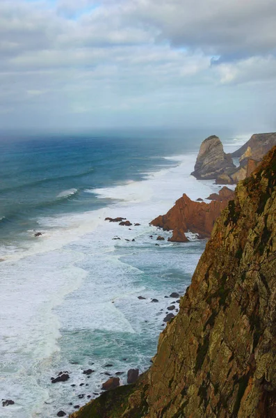 Stormy ocean with waves braking on the cliffs, cloudy sky background. Picturesque foggy morning landscape. Western point of Europe. Cape Roca (Cabo da Roca), Portugal. Travel and tourism concept.