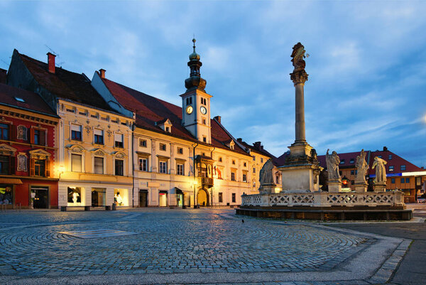 Wide-angle landscape view of illuminated Plague Column against cloudy sky. Ancient colorful building at the background. The Rotovz Town Hall Square in Maribor. Travel and tourism concept.