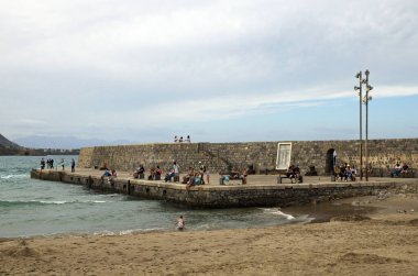 Cefalu, İtalya-28 Ekim 2023: Dinlenen insanlarla birlikte eski taş iskelenin manzarası. Bulutlu hava, Tyrhenian Denizi 'nde dalgalar. Seyahat ve turizm kavramı.